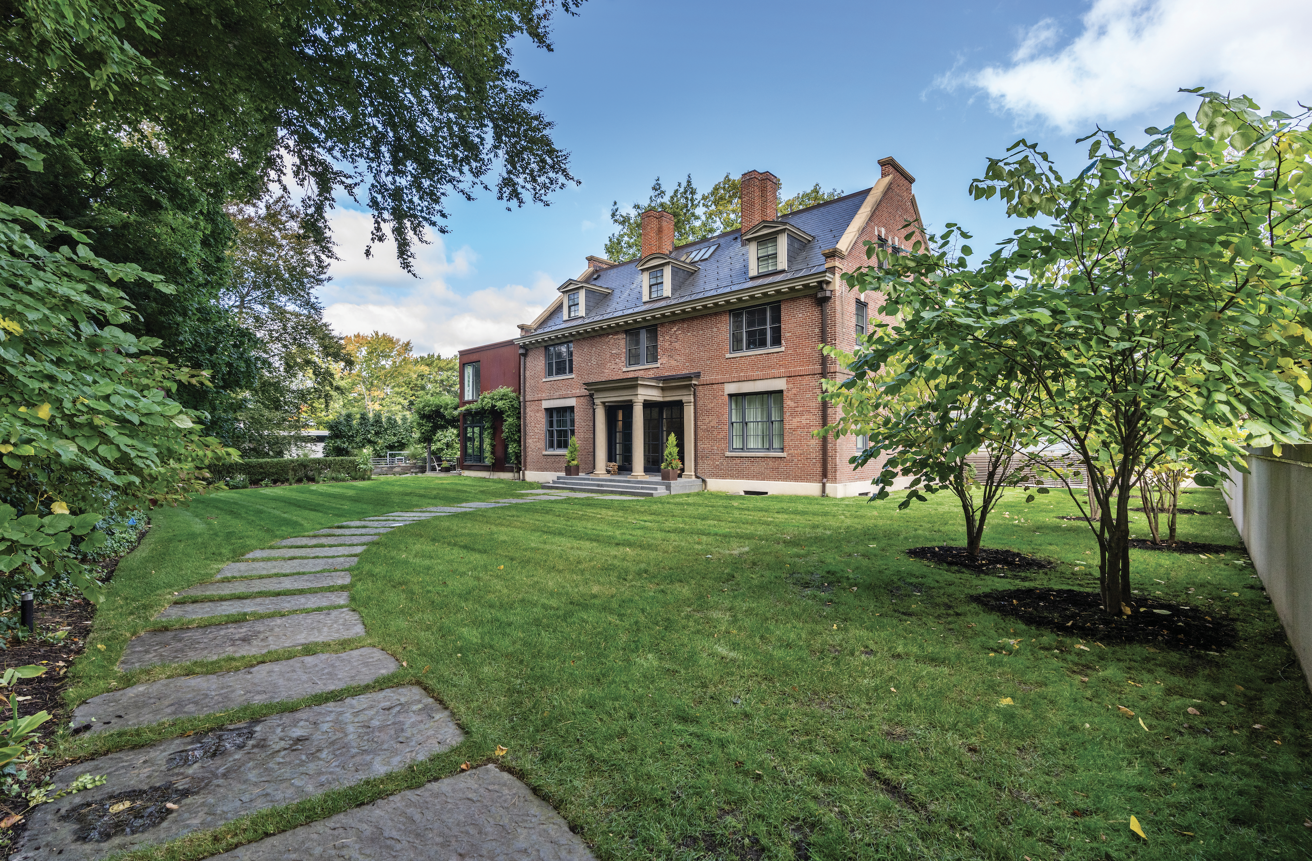 Historic red-brick home in West Cambridge surrounded by lush greenery and a stone walkway, showcasing timeless New England architecture and charm.